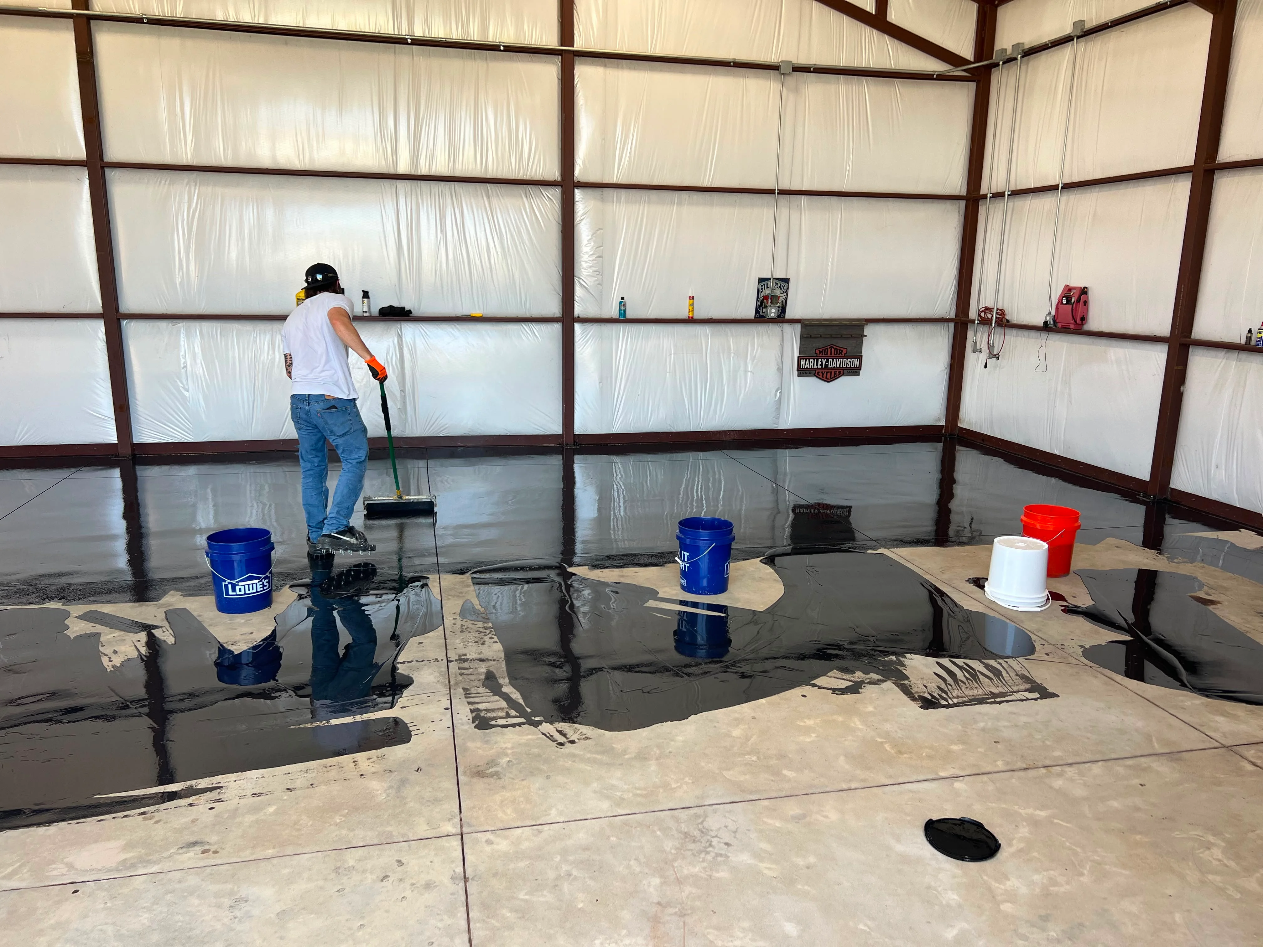Worker applying dark epoxy coating to garage floor with blue buckets and tools visible